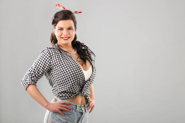Young woman posing in studio as a pin-up girl on gray background wearing tight shirt. About 25 years old, Caucasian plus-size brunette.